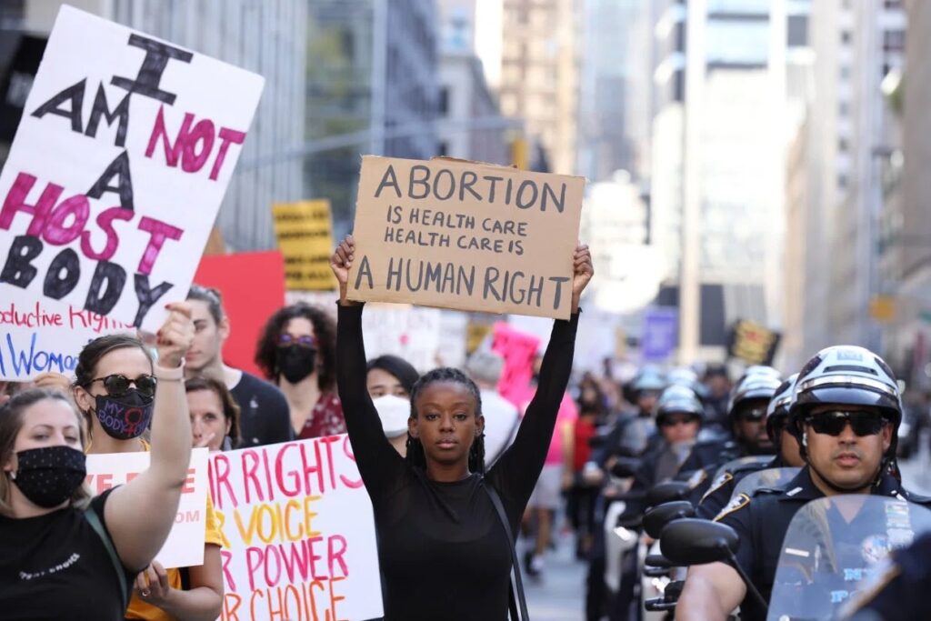 Pro-Abortion protesters march in New York City, 2021. Signs read "I am not a host body" and "abortion is healthcare and healthcare is a human right". Police are on motorcycles alongside protesters.